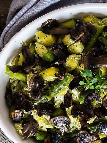 A white bowl filled with a salad of roasted mushrooms, diced potatoes, and green beans, garnished with a sprig of herbs, creates the perfect pair for a vegan pasta bake. Scattered cashews surround it alongside a spoon and rustic fabric on a wooden surface.
