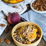A wooden bowl holds a dessert topped with chopped nuts, diced peaches, and a hint of Brie cheese with honey. A fresh peach and scattered pecans adorn the rustic wooden surface. White cloth napkins are draped delicately nearby.