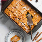 A rectangular baking dish filled with sliced pumpkin bread pudding, dusted with powdered sugar, evokes comforting flavors reminiscent of a recipe for carrot casserole. A serving spatula rests on top, with one piece on a patterned white plate nearby. Two cinnamon sticks adorn the marble surface.