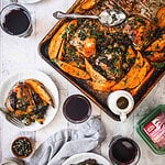 A Moroccan-inspired roasted chicken tray with sweet potatoes, olives, and herbs sits on a marble surface. Accompanying items include two glasses of red wine, a small dish of capers, a gravy boat, and a cloth napkin. A white plate holds a serving of this flavorful dish.