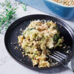 A serving of vegan broccoli casserole topped with breadcrumbs sits on a round, black plate. A fork rests beside the casserole. In the background, there is a blue baking dish and some vibrant green herbs, adding fresh allure to this plant-based delight.