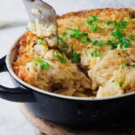 A casserole dish filled with creamy hashbrown chicken bake garnished with chopped green onions. A spatula lifts a portion, revealing the golden-brown crust and savory interior. The dish rests invitingly on a wooden board.