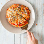 A person is about to serve a slice of a small, round breakfast pizza on a plate. The pizza, topped with melted cheese, tomato sauce, and herbs, sits invitingly on a textured white plate against a wooden surface background.