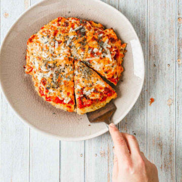 A person is about to serve a slice of a small, round breakfast pizza on a plate. The pizza, topped with melted cheese, tomato sauce, and herbs, sits invitingly on a textured white plate against a wooden surface background.