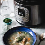 A bowl of steaming fish soup with vibrant vegetables and herbs sits invitingly in front of an Instant Pot on the kitchen counter. In the background, a small bowl of chopped herbs and a glass bottle complement the scene, while a spoon rests beside the hearty meal.