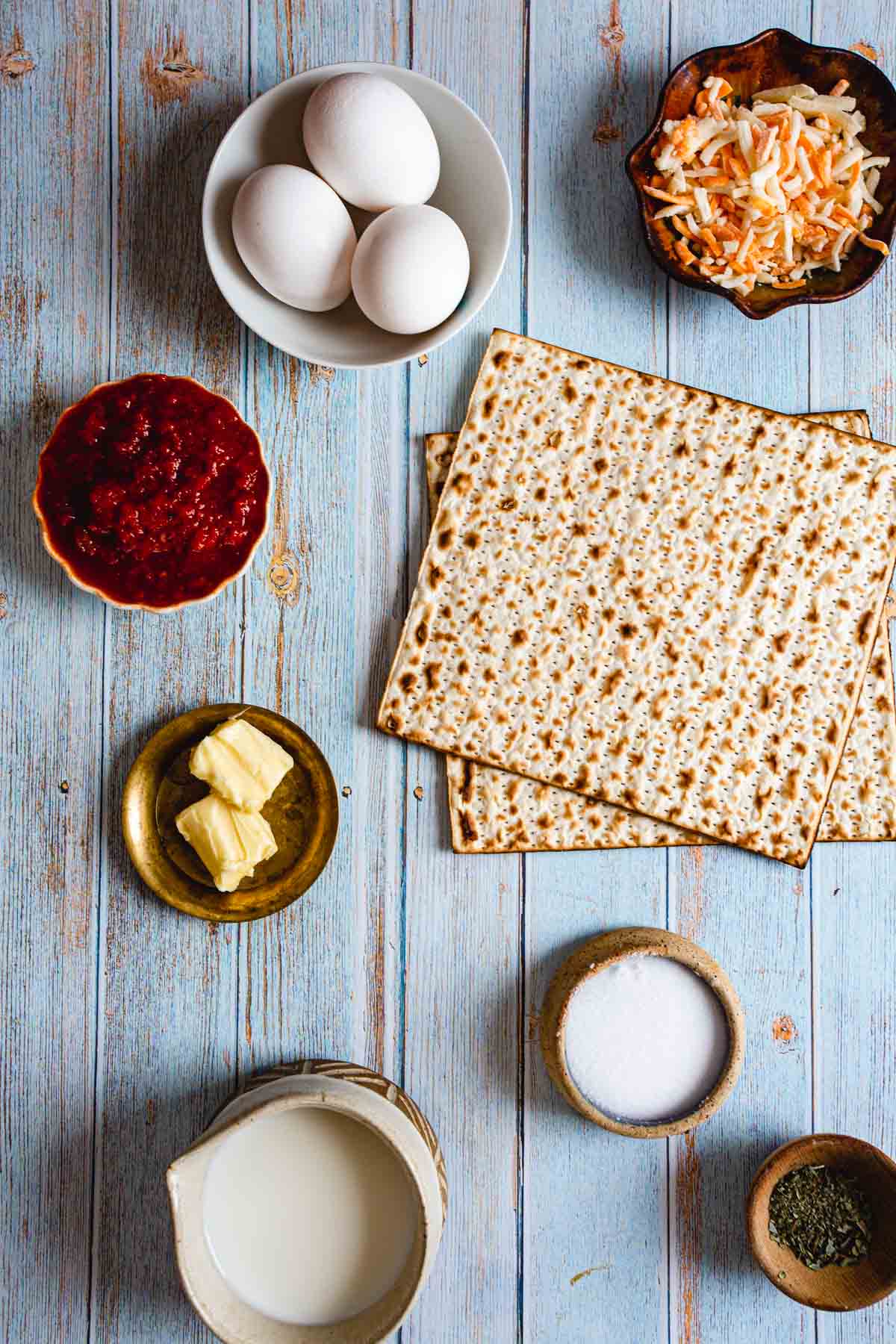Ingredients for how to make a breakfast pizza are displayed on a wooden surface: three eggs in a bowl, matzah, shredded vegetables in a bowl, tomato sauce, butter on a small plate, milk in a jug, salt, and herbs.