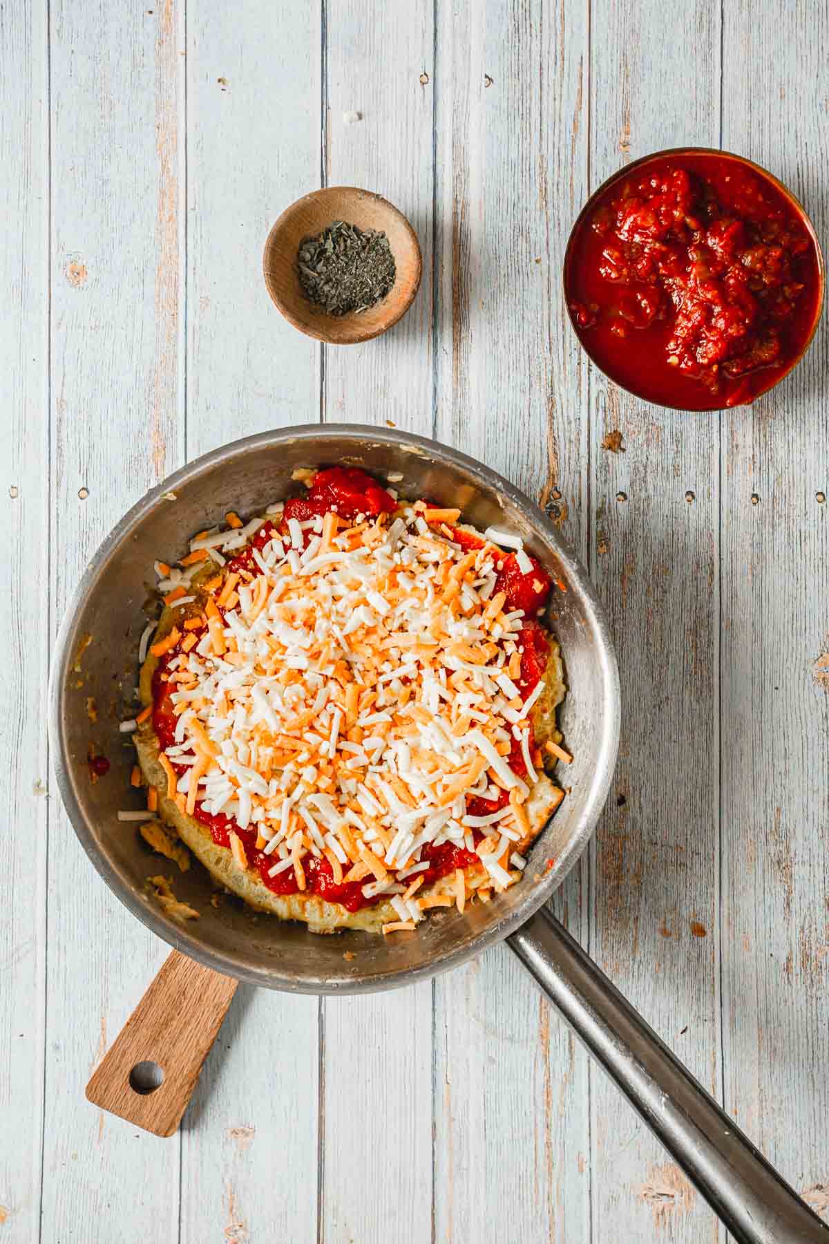 A skillet on a wooden table holds a breakfast pizza topped with mixed shredded cheese. Nearby, two small bowls await—one with chopped tomatoes and another with ground herbs—perfect for customizing your morning delight.