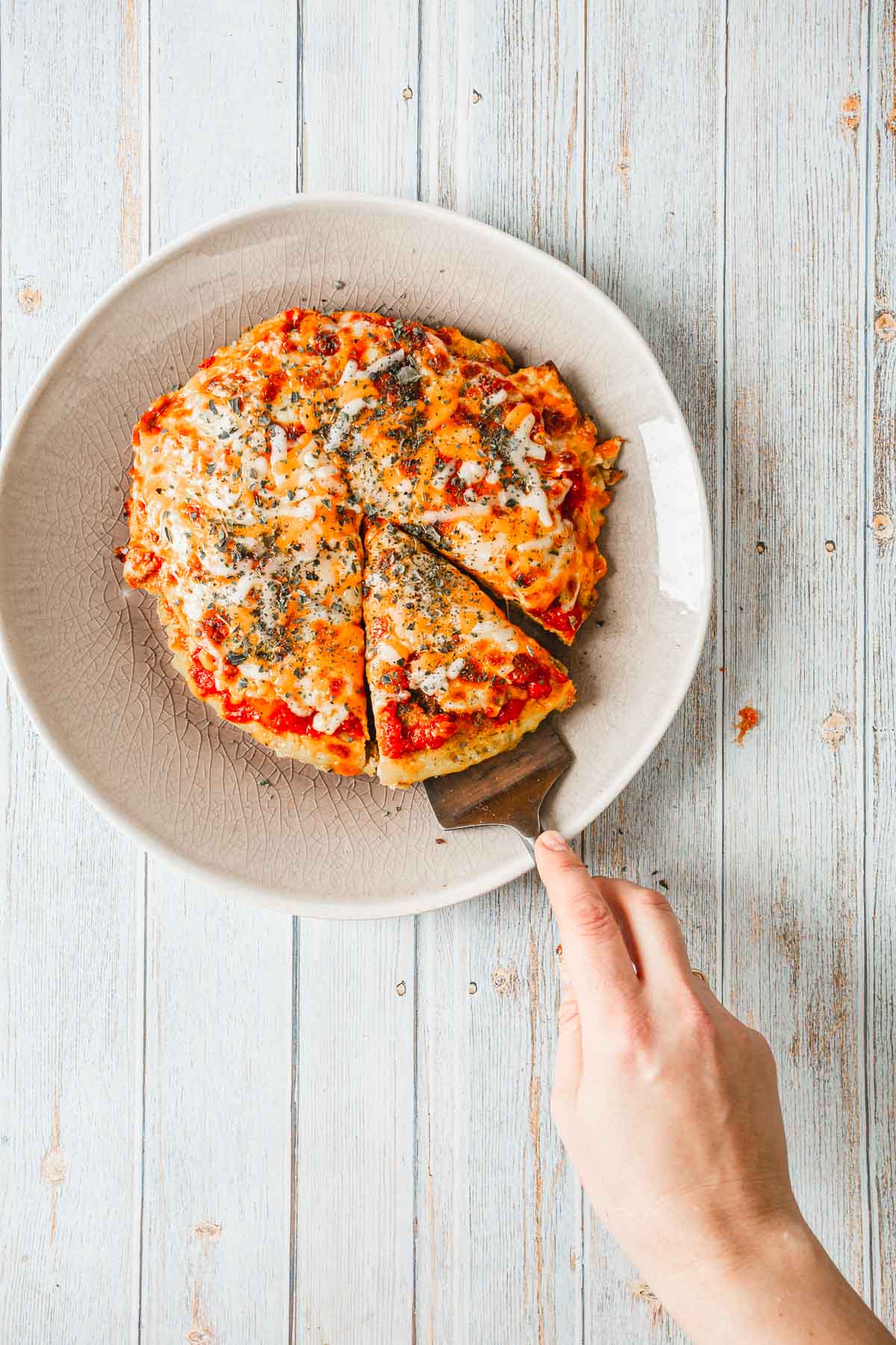 A hand is holding a spatula, expertly lifting a slice from a breakfast pizza on a white plate. The pizza, adorned with melted cheese and herbs, looks delectable atop the light-colored wooden surface.