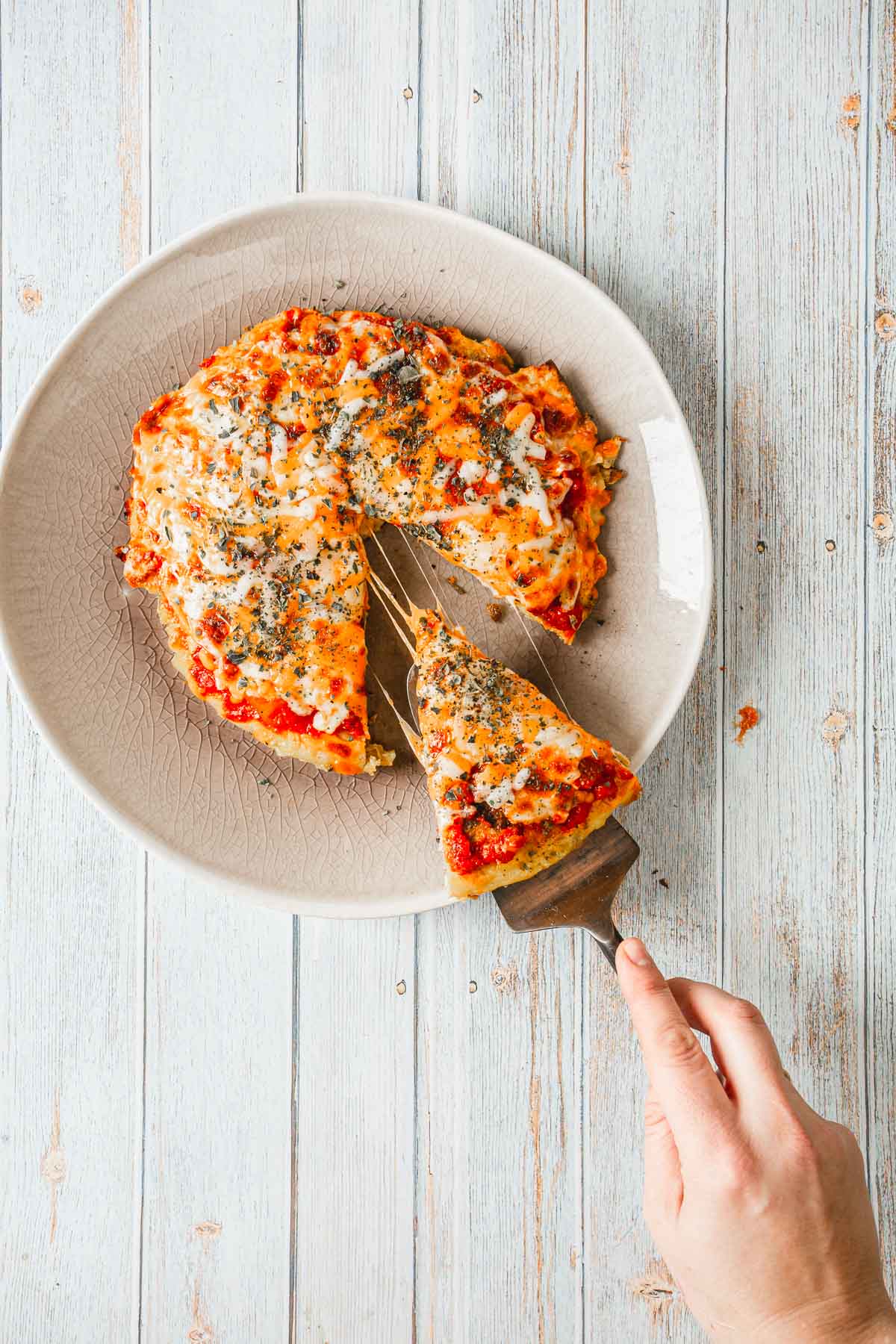 A hand holds a triangular slice of pizza above a textured beige plate with the remaining breakfast pizza. Topped with cheese, tomato sauce, and herbs, this delightful creation is perfect for any morning. The background features a light wooden surface, setting the stage for your culinary adventure.