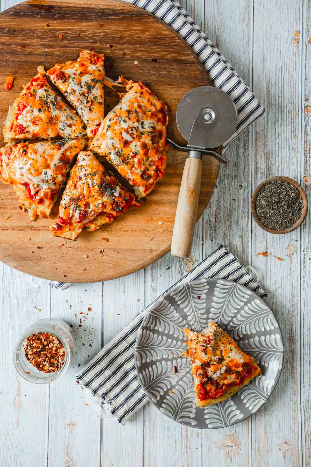 A pizza, cut into six slices, sits on a round wooden board beside a pizza cutter. A single slice rests on a patterned plate atop a striped napkin. Nearby, small bowls of crushed red pepper and dried herbs hint at the flavors behind how to make a breakfast pizza. The background is light wood.