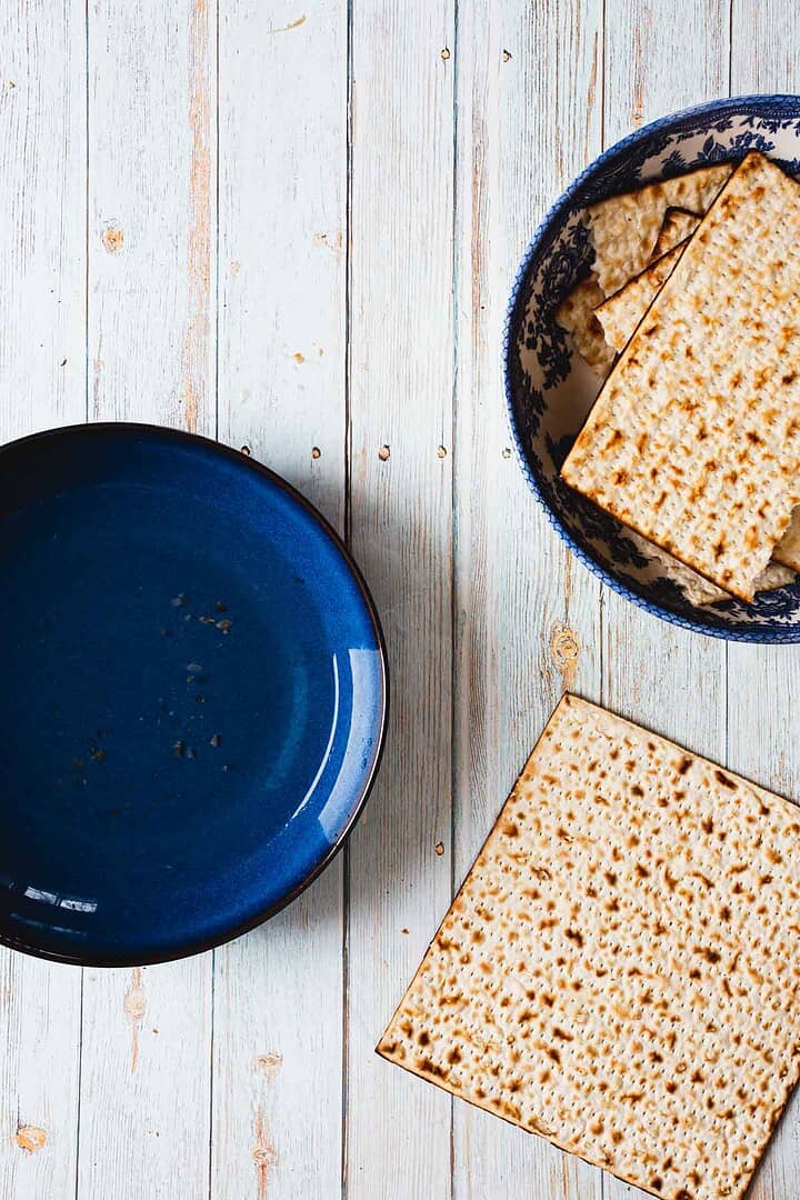 A blue bowl rests on a light wooden table. Beside it, a patterned bowl holds matzo crackers, one peeking out, crumbs scattered—a simple setup that could easily transform into the perfect backdrop for exploring *how to make a breakfast pizza*, merging rustic vibes with culinary creativity.
