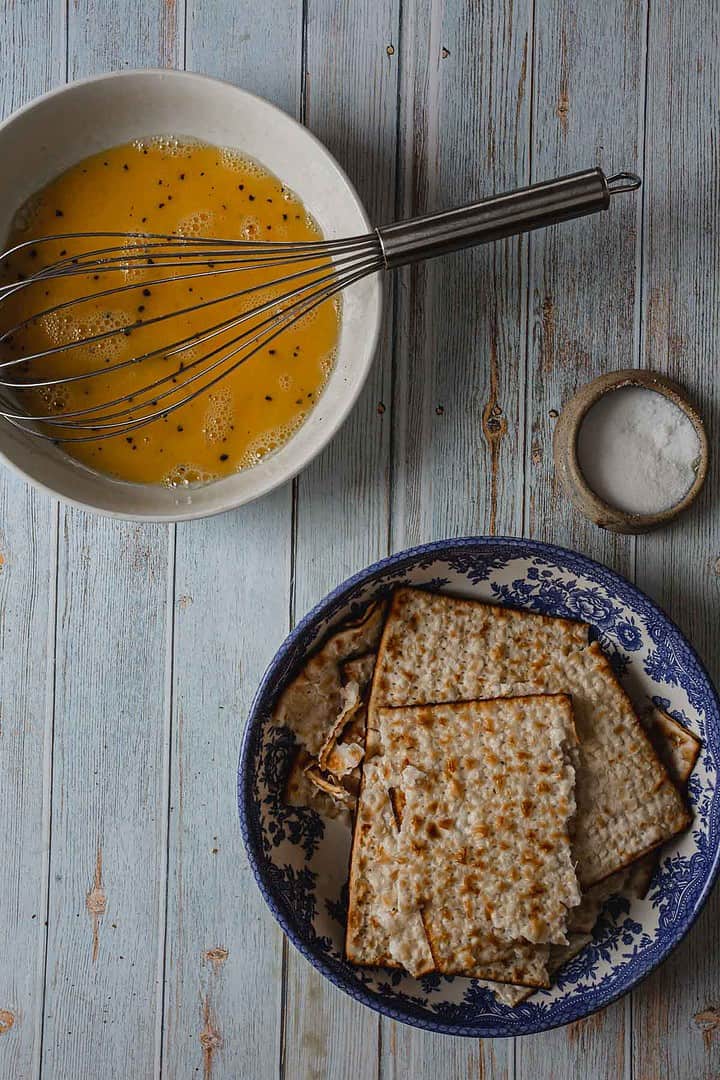 A bowl of beaten eggs with a whisk sits next to a small dish of salt on a light wooden surface, ready for the next step in how to make a breakfast pizza. Below it, a blue-patterned bowl holds several pieces of matzo.