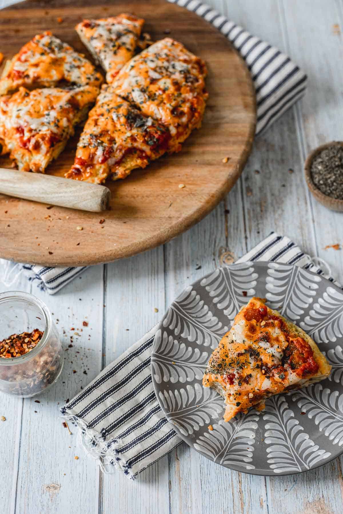 A wooden board with sliced cheese pizza topped with herbs showcases how to make a breakfast pizza. On a light wooden table, a single slice rests on a decorative gray plate with a white leaf pattern. Nearby, a small jar of red pepper flakes sits on a striped cloth.
