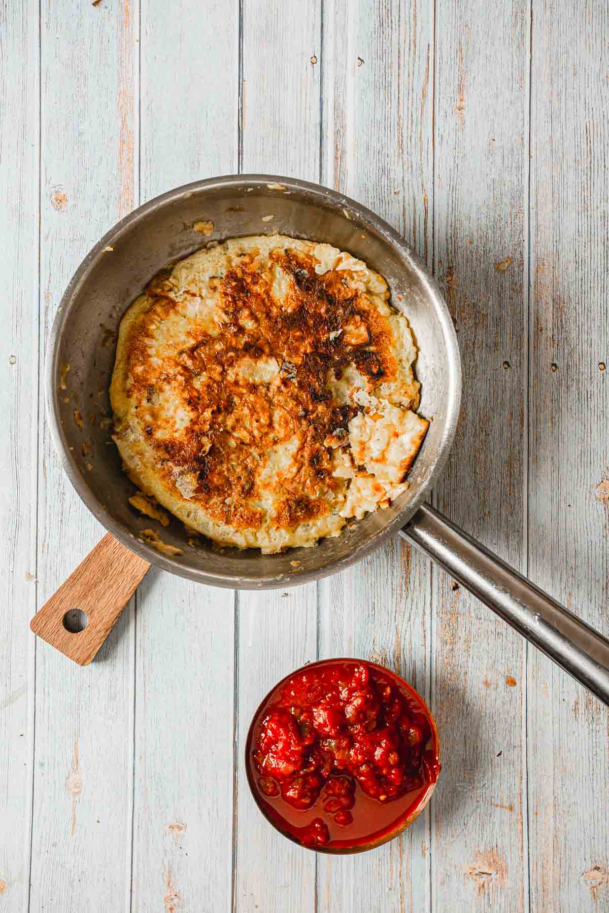 A pan with a large, browned potato pancake sits on a wooden surface next to a small bowl filled with red lingonberries. Perfect for those learning how to make a breakfast pizza, it's hearty and satisfying enough to start your day deliciously.