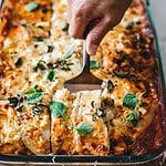 A hand uses a spatula to lift a slice of lasagna from the baking dish, resembling an easy moussaka recipe. The lasagna is topped with melted cheese and garnished with fresh herbs, appearing rich and savory, with visible layers and a golden-brown crust.