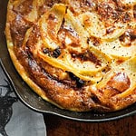 A close-up image of a golden-brown apple pie in a rustic metal pan. The pie, akin to a recipe for rutabaga casserole, is topped with thinly sliced apples and baked to crisp caramelization. It rests on a table, partially covering a black and white fabric.