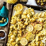 A baking sheet with roasted cauliflower, lemon slices, and olives accompanies a small decorative tagine and a bowl with more olives. Nearby, a bowl of dipping sauce complements the setup with a vibrant yellow flower, creating an appetizing prelude to your salmon casserole delight.