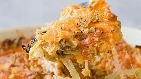 A close-up of a cheesy cabbage casserole being scooped from the dish. This golden-brown delight, made without canned soup, features a crunchy cracker topping and melted cheese. Strings of cheese stretch invitingly from the serving utensil to the dish.