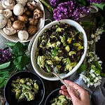 A hand holding a fork leans over a bowl of pasta with creamy sauce and mushrooms, reminiscent of a comforting vegan pasta bake. Surrounding the bowl are fresh mushrooms, small dishes with more pasta, and lilac flowers adding a touch of elegance.
