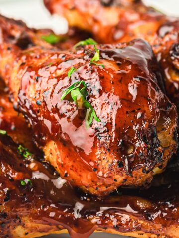 A plate of glazed barbecue chicken drumsticks garnished with small parsley leaves. The chicken is coated in a shiny, dark sauce, suggesting a rich, flavorful marinade. The dish is arranged to highlight the crispy, caramelized skin.