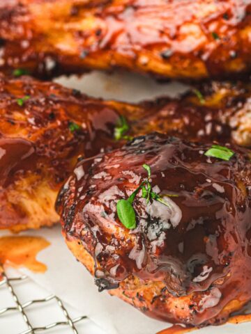 Close-up of glazed barbecue chicken legs on a cooling rack, garnished with fresh herbs. The chicken is coated in a rich, glossy sauce, with small sprigs of greenery scattered around, resting on a white parchment paper.