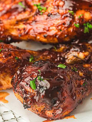 Close-up of three grilled chicken drumsticks coated in barbecue sauce, garnished with fresh herbs. They are placed on a cooling rack lined with parchment paper. Fresh greenery is visible in the foreground.