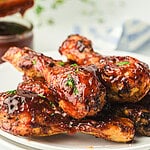 Close-up of barbecue-glazed chicken drumsticks stacked on a white plate. The drumsticks are coated with a glossy sauce and garnished with small pieces of green herbs. In the background, there's a blurred view of a sauce container and a blue-striped napkin.