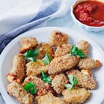 A white plate with golden-brown breaded chicken nuggets and crispy air fryer cheese bites, garnished with fresh parsley leaves. A bowl of red tomato sauce is in the background, partially wrapped in a gray and white cloth.