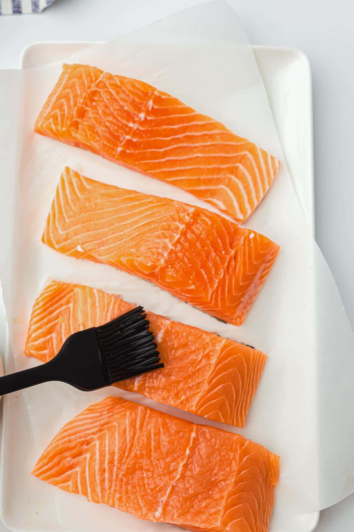 Four raw salmon fillets rest on a white rectangular tray lined with parchment paper, ready for transformation into air fryer salmon. A black brush sits nearby, hinting at seasoning or marination. The backdrop remains light and unobtrusive, keeping attention on the culinary preparation.