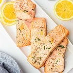 A white rectangular plate features air fryer salmon fillets, garnished with chopped herbs and cracked pepper. Lemon slices are artfully placed around the salmon. A blue-gray cloth is partially visible in the bottom left corner, adding a touch of elegance to the presentation.