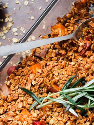 A baking dish filled with baked oatmeal topped with fresh rosemary sprigs. A spoon rests inside the dish. Dried oats and cranberries are scattered on the wooden table, and a small bowl of cranberries is visible in the background.