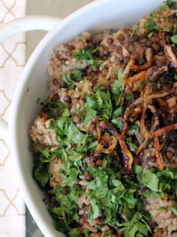 A dish of mujadara is served in a white bowl. The dish consists of rice and lentils topped with fresh parsley and crispy fried onions. A wooden spoon lies next to the bowl on a patterned cloth.
