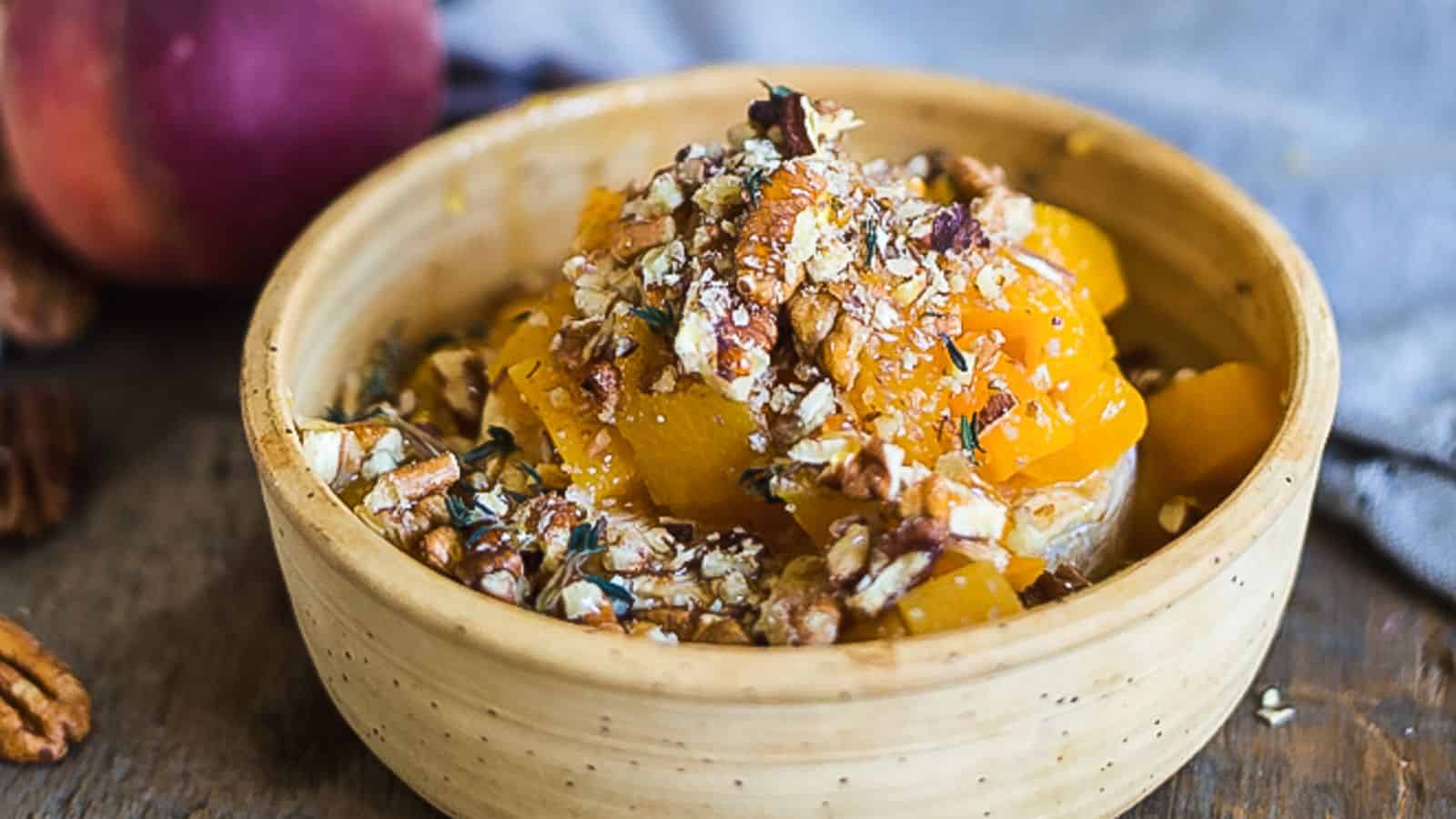 A wooden bowl filled with melted cheese, chopped peaches, and pecans. A bread slice is seen in the background, and a piece of bread is used to scoop up the cheesy mixture. Pecans are scattered around the bowl.