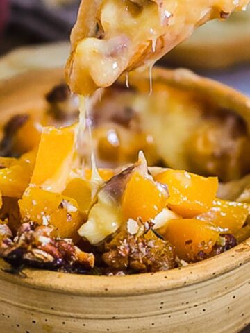 A wooden bowl filled with melted cheese, chopped peaches, and pecans. A bread slice is seen in the background, and a piece of bread is used to scoop up the cheesy mixture. Pecans are scattered around the bowl.