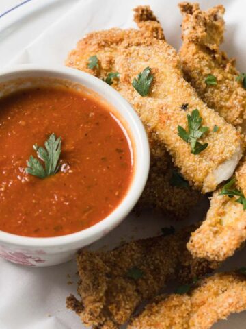 Breaded chicken tenders on a white plate with a bowl of marinara sauce, garnished with parsley—perfect for quick and cozy meals.