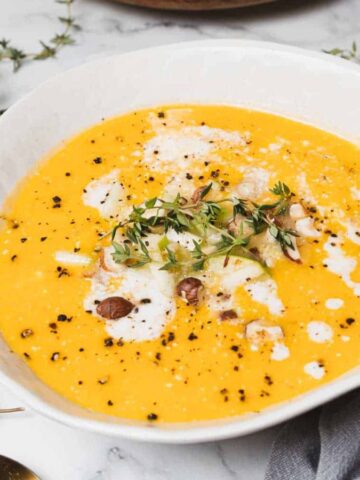 A bowl of creamy orange soup garnished with herbs, chopped nuts, and drizzled cream sits on a marble surface. A small bowl of nuts and sprigs of fresh herbs are nearby, alongside a gray napkin.
