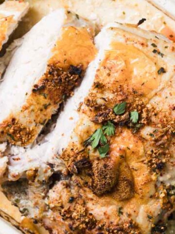 A roasted turkey breast is sliced and displayed in a baking dish. The surface is seasoned with herbs and spices. Accompanying it are vegetables like green beans, corn, and onions, all cooked together.