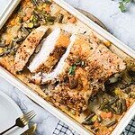 A rectangular dish contains Cajun-style baked turkey breast on a bed of mixed vegetables, including green beans, carrots, and corn. The meat is garnished, and there is a fork and knife next to the dish on a white plate. A sprig of herbs is visible on top.