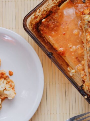 A baked dish with a large serving of cheesy cauliflower in a glass baking dish is next to a white plate holding a portion of the same dish. The background is a light wooden surface. A fork is visible beside the plate.
