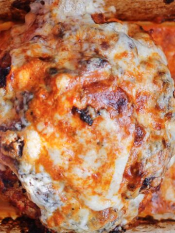 Baked whole cauliflower in a glass dish, covered with melted cheese and a creamy, reddish sauce, browned on the edges. The dish shows burnt and crispy spots around the cauliflower.