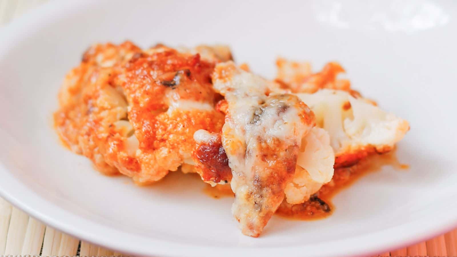 A baked dish with a large serving of cheesy cauliflower in a glass baking dish is next to a white plate holding a portion of the same dish. The background is a light wooden surface. A fork is visible beside the plate.