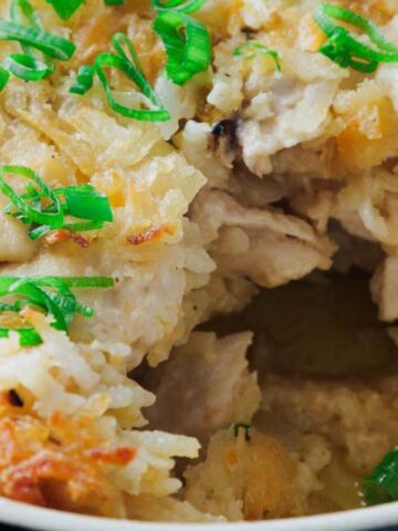 A close-up of a baked dish topped with grated cheese or potatoes and garnished with chopped green onions. A portion is removed, revealing a filling that appears to include pieces of meat. The dish is in a black, round container.