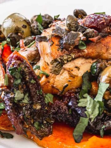 A plate of roasted vegetables and chicken is garnished with capers and fresh herbs. The dish includes sweet potatoes, olives, and mushrooms, and is accompanied by a fork.