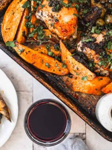 Roasted chicken with sweet potato wedges, herbs, and olives on a tray, served with wine and sauce—perfect for those who crave hearty Comfort Meals.