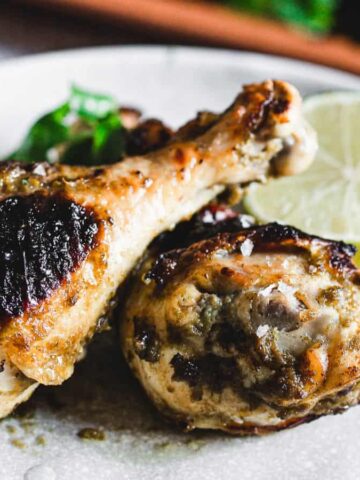 A plate featuring two grilled chicken drumsticks, one with a charred top. A slice of lime and some greens accompany the chicken, all on a gray speckled plate.