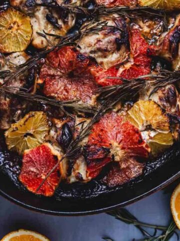 A roasted dish featuring chicken pieces garnished with slices of oranges and grapefruits, topped with sprigs of rosemary, in an oval orange pan. Slices of citrus fruits are scattered around on a blue surface.