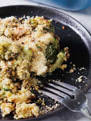 A black plate with a serving of broccoli casserole, topped with breadcrumbs. A fork rests on the plate. The background is a marble surface, with some green plant decorations partially visible.