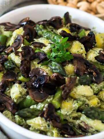 A bowl of pasta salad featuring mushrooms, asparagus, and a creamy green sauce. The dish is garnished with fresh herbs. A small bowl of nuts and some greenery are in the blurred background.