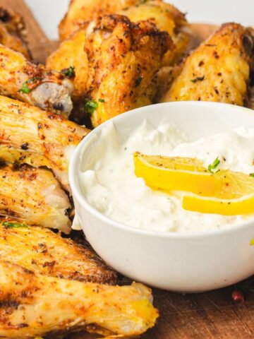 A wooden board with seasoned chicken wings, a bowl of creamy dipping sauce topped with lemon slices, and carrot and celery sticks on the side.