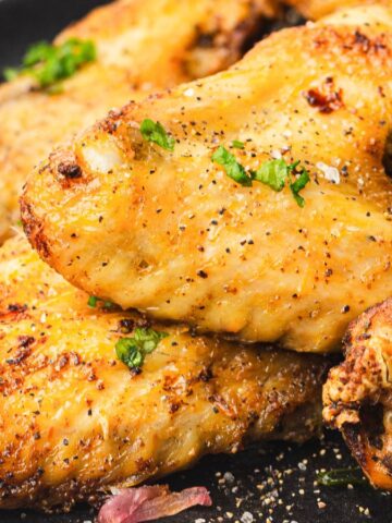 Close-up of a plate of seasoned and grilled chicken wings garnished with herbs. Lemon wedges are visible in the background, adding a hint of color. The wings appear crispy and well-cooked, resting on a dark plate.